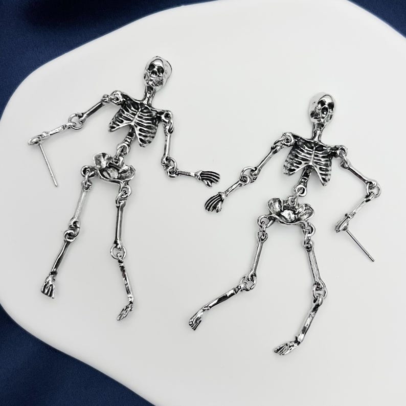 May include: A pair of silver-toned metal earrings shaped like articulated human skeletons. Each earring features a detailed skull, rib cage, pelvic bone, and jointed limbs connected by small metal rings, with a post backing for attachment.