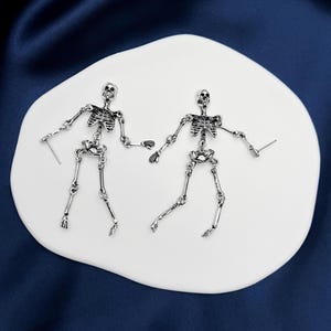 May include: A pair of silver-toned metal earrings shaped like articulated human skeletons. Each earring features a detailed skull, rib cage, pelvic bone, and jointed limbs connected by small metal rings, with a post backing for pierced ears.