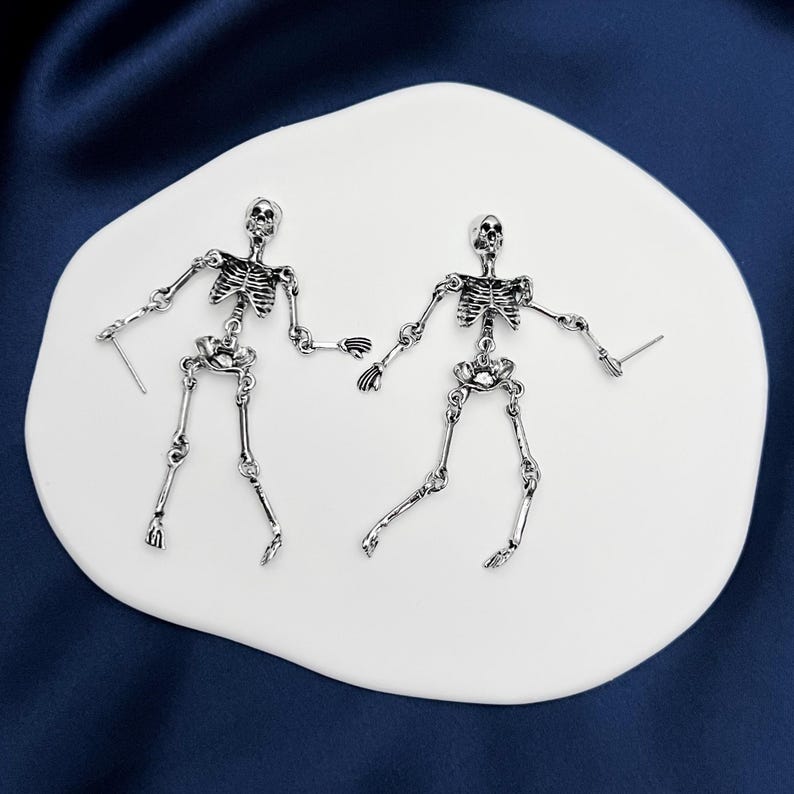 May include: A pair of silver-toned metal earrings shaped like articulated human skeletons. Each earring features a detailed skull, rib cage, pelvic bone, and jointed limbs connected by small metal rings, with a post backing for pierced ears.