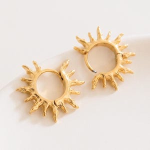 Op de afbeelding: Een paar goudkleurige oorringen met een zonnestraalontwerp. Elke cirkelvormige ring is versierd met stralende, getextureerde punten die het uiterlijk van zonnestralen nabootsen. De oorbellen hebben een gepolijste metalen afwerking en een scharniersluiting.