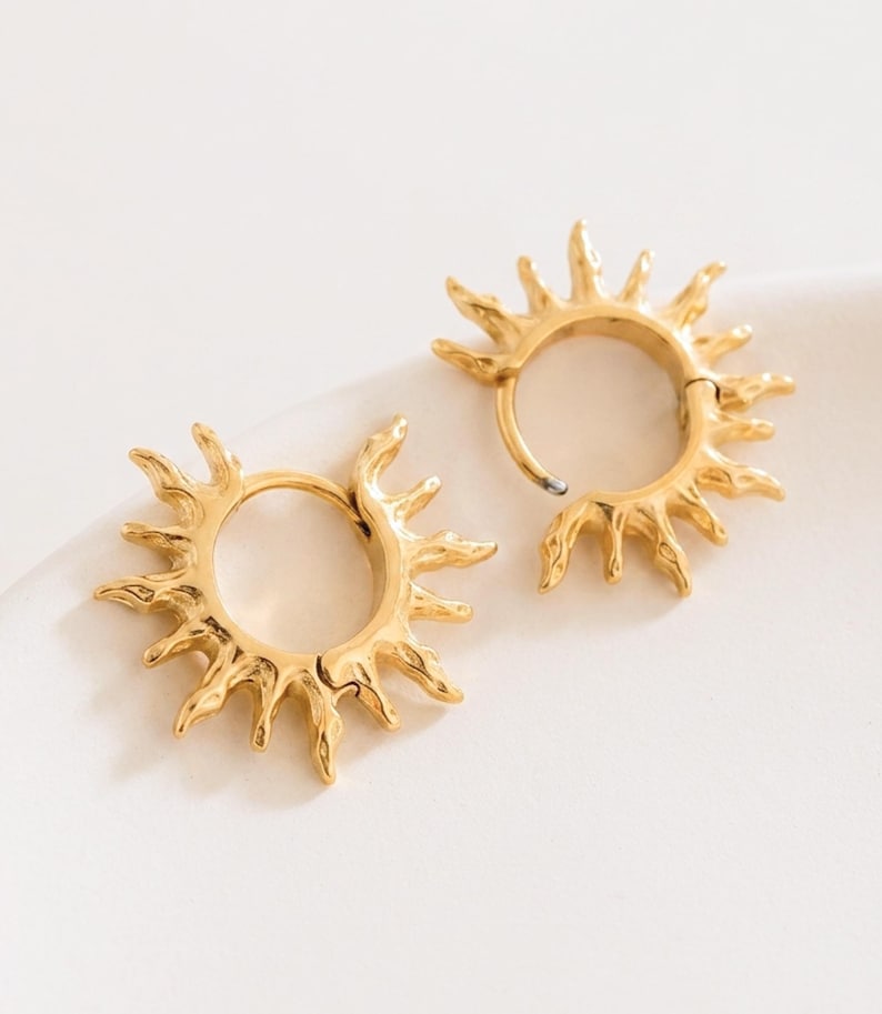 Op de afbeelding: Een paar goudkleurige oorringen met een zonnestraalontwerp. Elke cirkelvormige ring is versierd met stralende, getextureerde punten die het uiterlijk van zonnestralen nabootsen. De oorbellen hebben een gepolijste metalen afwerking en een scharniersluiting.