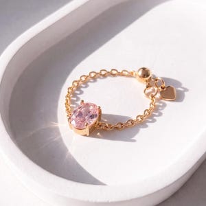 May include: A gold chain ring featuring an oval-cut light pink gemstone set in a four-prong setting. The ring includes a small dangling heart charm and a round gold bead, creating a flexible band design with a polished metallic finish.