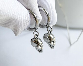 May include: A pair of silver-toned hoop earrings featuring small, detailed skull charms hanging from each hoop. The earrings are crafted from a polished metallic material with a smooth finish, showcasing a realistic skull design with visible eye sockets and teeth.