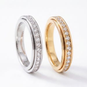 May include: Two rings standing upright side by side. One ring is made of polished silver-toned metal and the other is made of polished gold-toned metal. Both rings feature a central channel set with a continuous row of small, clear, round-cut stones.