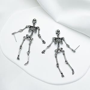 May include: A pair of silver-toned metal skeleton earrings featuring articulated limbs and a post-back closure. Each earring depicts a full human skeletal structure with detailed rib cages, skulls, and jointed arms and legs in a polished finish.