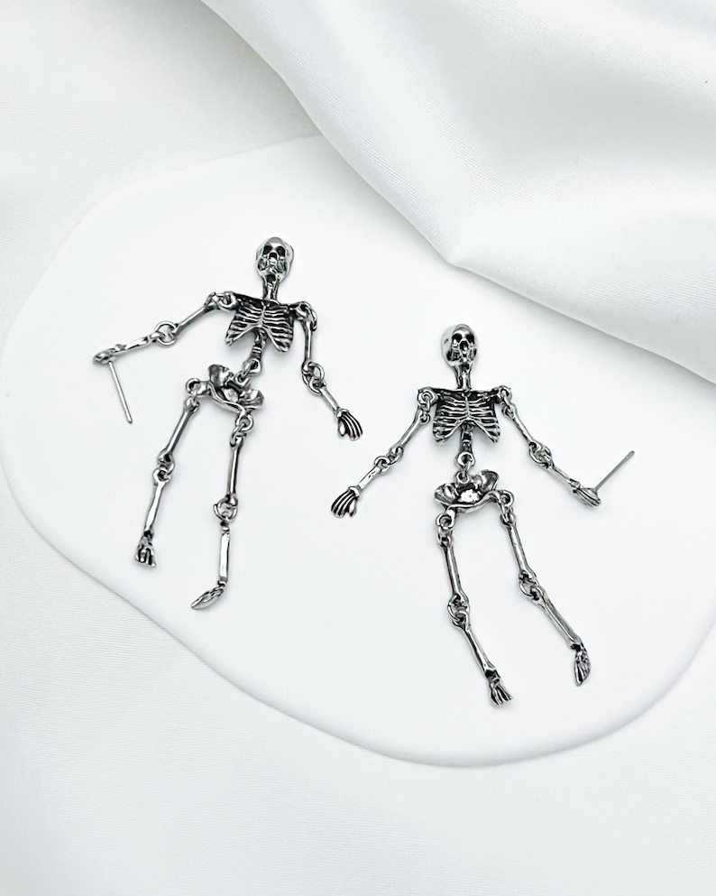 May include: A pair of silver-toned metal skeleton earrings featuring articulated limbs and a post-back closure. Each earring depicts a full human skeletal structure with detailed rib cages, skulls, and jointed arms and legs in a polished finish.