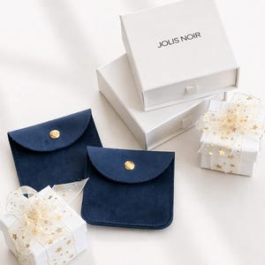 May include: An assortment of jewelry packaging, including two navy blue suede pouches with gold snap closures, white gift boxes, and a box with the text "JOLIS NOIR". The gift boxes are adorned with gold star ribbon.