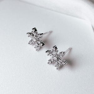 Op de afbeelding: Een paar zilverkleurige sneeuwvlok oorknopjes. Elke oorbel heeft een gedetailleerd sneeuwvlokontwerp met meerdere punten en kleine, heldere accenten. De oorbellen staan tegen een witte achtergrond.