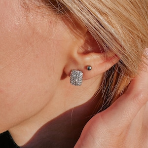 May include: A rectangular stud earring featuring multiple rows of clear, sparkling stones set in a metallic frame. The earring is positioned on an earlobe, accompanied by a small, simple silver-toned ball stud earring located just above it.