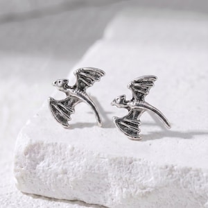 May include: A pair of silver-colored dragon-shaped stud earrings. Each earring features a detailed dragon with outstretched wings and a curved tail. The earrings are displayed on a textured white surface.