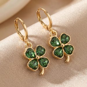 Peut inclure: Une paire de boucles d'oreilles cr&eacute;oles dor&eacute;es avec des pendentifs en forme de tr&egrave;fle &agrave; quatre feuilles. Chaque tr&egrave;fle est compos&eacute; de quatre cristaux vert fonc&eacute; facett&eacute;s sertis dans une monture en m&eacute;tal dor&eacute;, suspendus &agrave; une attache cr&eacute;ole.