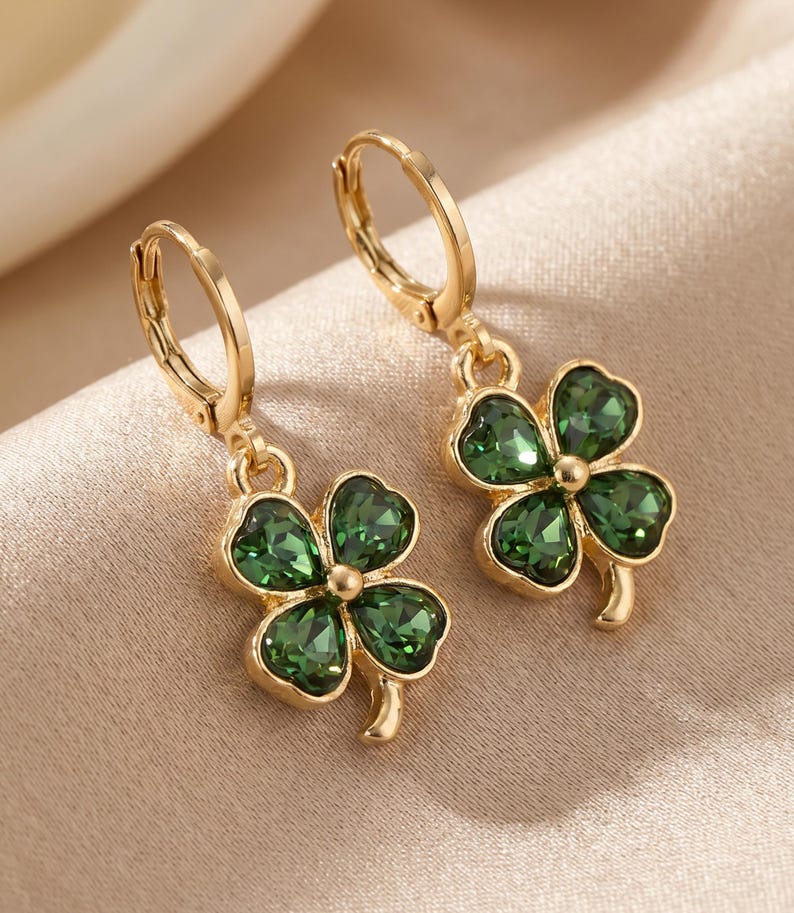 Peut inclure: Une paire de boucles d'oreilles cr&eacute;oles dor&eacute;es avec des pendentifs en forme de tr&egrave;fle &agrave; quatre feuilles. Chaque tr&egrave;fle est compos&eacute; de quatre cristaux vert fonc&eacute; facett&eacute;s sertis dans une monture en m&eacute;tal dor&eacute;, suspendus &agrave; une attache cr&eacute;ole.