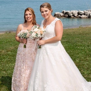 May include: Two people in wedding attire stand on a grassy area near a body of water. The person on the left is wearing a blush-colored dress with floral embroidery. The person on the right is wearing a white lace wedding dress. Both people are holding bouquets of flowers.