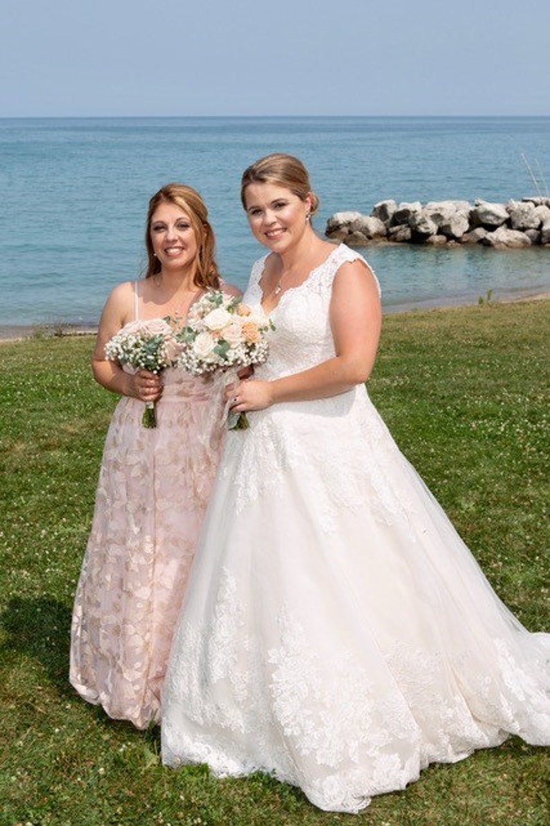 May include: Two people in wedding attire stand on a grassy area near a body of water. The person on the left is wearing a blush-colored dress with floral embroidery. The person on the right is wearing a white lace wedding dress. Both people are holding bouquets of flowers.