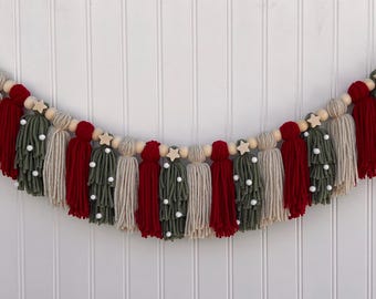 Puede incluir: Una guirnalda navideña festiva con borlas de hilo rojo, verde y crema, cuentas de madera y detalles de estrellas blancas.