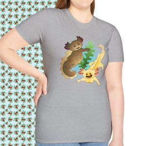 Könnte beinhalten: Hellgraues T-Shirt mit einer farbenfrohen Illustration von zwei Axolotln. Einer ist braun, der andere gelb, beide lächeln. Sie sind vor einem Hintergrund aus Sand und Wasserpflanzen platziert.
