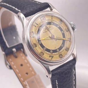 May include: Vintage Mido Multifort wristwatch with a silver-tone case, a gold-tone dial, and black leather band. The watch features a textured dial with Arabic numerals and a date display. The black leather band has white stitching.