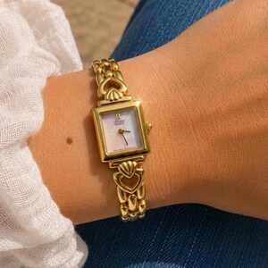 May include: Gold Citizen Quartz watch with a square face and a mother-of-pearl dial. The watch features a gold link band with heart-shaped accents. The watch is approximately 1.9 cm wide.