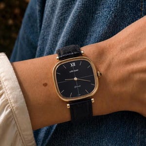 Peut inclure: Une montre-bracelet carrée dorée avec un cadran noir, portant les inscriptions Wittnauer et Geneve. La montre possède de fines aiguilles dorées, un chiffre romain XII en haut et un bracelet en cuir noir texturé façon crocodile avec des attaches dorées.