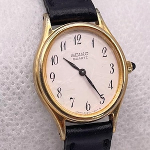 Può includere: Un orologio da polso Seiko Quartz color oro con quadrante bianco e lancette nere. L'orologio ha un cinturino in pelle nera. Il quadrante mostra i numeri da 1 a 12. L'orologio è un design classico ed elegante.