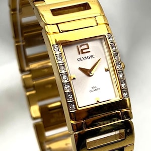 May include: Gold-toned Olympic quartz watch with a rectangular face and a gold link band. The watch face is white with the number 12 in brown, and the word "OLYMPIC" is printed on the face. The watch is adorned with small crystals.