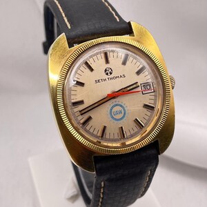 May include: A vintage Seth Thomas wristwatch with a gold-toned case and a cream-colored face. The watch features black hands, a red second hand, and a date display. The black leather band is stitched with white thread.