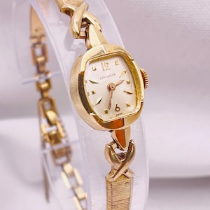 May include: A gold-tone Longines wristwatch featuring a small, rounded rectangular case with a cream-coloured dial, gold-tone hands, and numerical markers at 6 and 12. The watch has a textured metal link bracelet with decorative X-shaped connectors.