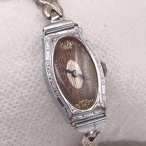 May include: A vintage silver-toned wristwatch with an oval face and a brown dial. The watch features ornate detailing around the face and on the band. The watch face displays the numbers 1 through 12. The band is made of silver-toned metal.