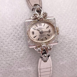 May include: A vintage Waltham Incabloc wristwatch with a silver rectangular face and a matching strap. The watch face displays the numbers 12, 3, 6, and 9, with fine hands and the brand name. The strap has a textured design.