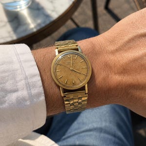 May include: A gold-toned Bulova Accutron watch with a matching gold band. The watch face is a light gold color with gold hour markers and hands. The watch is on a wrist, with a white shirt and blue jeans visible.