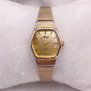 Peut inclure: Une montre à quartz Marquis dorée avec un cadran rectangulaire et un bracelet en maille dorée. Le cadran est doré avec des index et des aiguilles noirs. Le mot "Quartz" est imprimé sur le cadran.