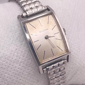 May include: A vintage Bulova wristwatch with a rectangular, gold-toned face and silver-toned band. The watch face features black stick hour markers and the word "Bulova" printed in black. The band is a flexible, woven design.