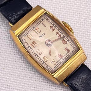 Könnte beinhalten: Eine vintage, goldfarbene Oris Armbanduhr mit rechteckigem Zifferblatt und schwarzem Lederarmband. Das Zifferblatt zeigt goldfarbene Ziffern und Zeiger, mit den Worten "ORIS" und "SWISS MADE". Eine klassische Uhr.