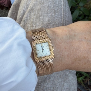 Könnte beinhalten: Eine goldfarbene Armbanduhr mit rechteckigem Zifferblatt und Mesh-Armband. Das Zifferblatt ist weiß mit schwarzen Zeigern und Markierungen. Die Uhr wird am Handgelenk getragen, mit einem weißen Hemd und einer beigen Hose im Hintergrund.