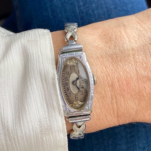May include: A vintage silver-coloured wristwatch with an oval face and a light-coloured dial. The watch has black numerals and hands, and is attached to a silver-coloured band with decorative links. The watch is worn on a wrist.