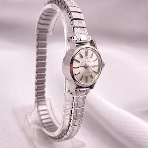 May include: A vintage silver-toned wristwatch with a silver band. The watch face is a light cream color with silver hands and markers. The watch is displayed on a clear stand. The watch face reads "ETERNA-MATIC" and "GOLDEN HEART".