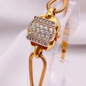 May include: A gold-tone wristwatch featuring a rectangular case with a silver-tone face encrusted with clear round rhinestones. The watch has a flexible, snake-style gold-tone metal band and a small winding crown on the right side of the case.
