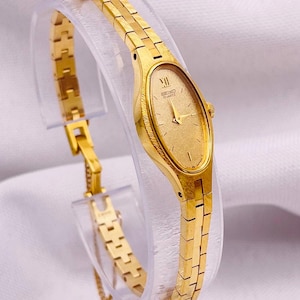 Può includere: Un orologio Seiko Quartz color oro con cassa ovale e quadrante dorato testurizzato. L'orologio presenta un sottile cinturino in metallo a maglie e un numero romano in corrispondenza delle ore dodici. Il quadrante riporta la scritta Seiko Quartz.