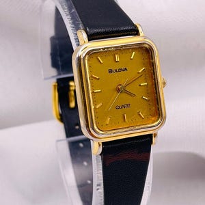 May include: A Bulova quartz wristwatch featuring a rectangular gold-tone case with a matching gold dial, gold-tone hour markers, and three hands. The watch is attached to a black leather strap with a gold-tone buckle and a visible dark red accent.