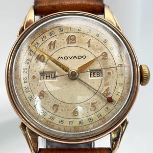 Może przedstawiać: Zegarek na rękę Movado w stylu vintage ze złotą kopertą i kremową tarczą. Zegarek posiada złote wskazówki, wyświetlacz dnia i daty oraz brązowy skórzany pasek. Tarcza zegarka wyświetla dzień tygodnia i datę.