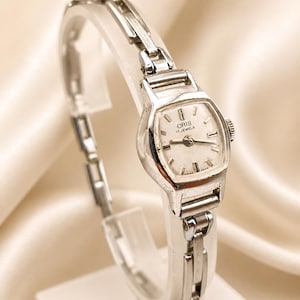 Könnte beinhalten: Eine silberfarbene Vintage-Armbanduhr mit rechteckigem Zifferblatt und Gliederarmband. Das Zifferblatt ist weiß mit schwarzen Ziffern und Zeigern. Die Uhr ist mit "ORIS 17 JEWELS SWISS MADE" gekennzeichnet.