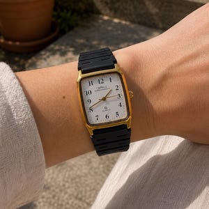 May include: Gold-coloured rectangular watch with a white face and black strap. The watch face displays the numbers 1 to 12, with the brand name "CAPRICE" and the word "QUARTZ" printed on the face. The black strap is ribbed.