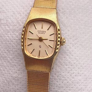 Könnte beinhalten: Eine goldfarbene Citizen Quartz Armbanduhr mit rechteckigem Zifferblatt und Mesh-Armband. Das Zifferblatt ist cremefarben mit goldenen Zeigern und Markierungen. Auf dem Zifferblatt steht "CITIZEN QUARTZ".