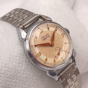 Op de afbeelding: Vintage Nivada Farren Price polshorloge met een zilverkleurige metalen band en kast. De wijzerplaat is lichtbeige met goudkleurige wijzers en cijfers. Een kleine secondenwijzer staat onderaan.