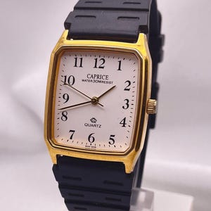 May include: A gold-coloured rectangular watch with a white face and black strap. The watch face displays the brand "CAPRICE", the word "QUARTZ", and the text "WATER 30M RESIST". The watch has black hands and a black rubber strap.
