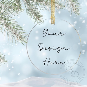 May include: A clear, round Christmas ornament with the text "Your Design Here" in black script. The ornament hangs from a gold ribbon against a snowy, blue background with frosted pine branches.