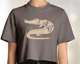Può includere: Un crop top grigio con un disegno di un alligatore dei cartoni animati e la parola "Later" incorporata nel suo corpo. L'alligatore è beige con accenti verdi. La maglietta ha maniche corte e girocollo.