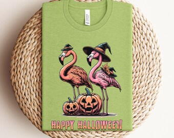 May include: A green t-shirt with a Halloween design. It features two pink flamingos wearing witch hats, standing beside carved pumpkins. The words "HAPPY HALLOWEEN" are printed below. A fun, seasonal design.