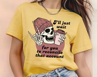 May include: Yellow short-sleeved t-shirt with a graphic of a skeleton wearing a pink beanie and holding a coffee cup. The text on the shirt reads "I'll just wait for you to reconcile that account."