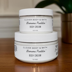 May include: Two white jars of body cream with black labels. The smaller jar is labeled "CLOVER BODY & BATH Banana Puddin' BODY CREAM 4 OZ (112 G)". The larger jar is labeled "CLOVER BODY & BATH Banana Puddin' BODY CREAM 7 OZ (196 G)".